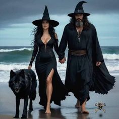 two people dressed in witches walking on the beach with a black dog next to them