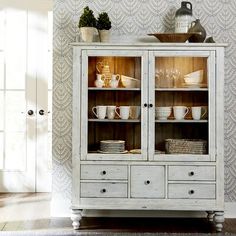 a white china cabinet with glass doors and dishes on it's sides, in front of a wallpapered background