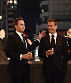 two men in tuxedos standing next to each other at a bar with people behind them