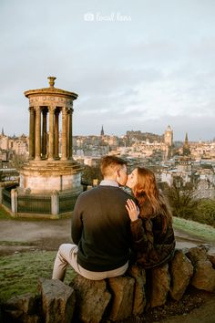 a man and woman sitting on top of a stone wall next to each other in front of a city