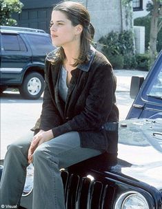 a woman sitting on the hood of a car in front of a parked vehicle and looking off into the distance