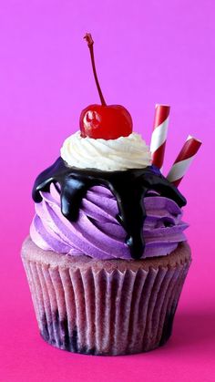 a cupcake with purple frosting and a cherry on top, sitting on a pink surface