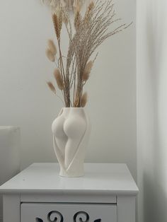 a white vase filled with dried flowers on top of a dresser next to a wall