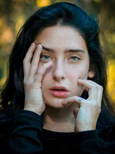 a woman with long black hair holding her hand to her face and looking at the camera