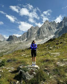 a woman standing on top of a lush green field next to mountains with clouds in the sky