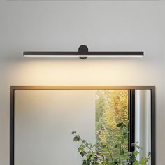 a bathroom mirror sitting above a plant in front of a wall mounted light on top of a counter