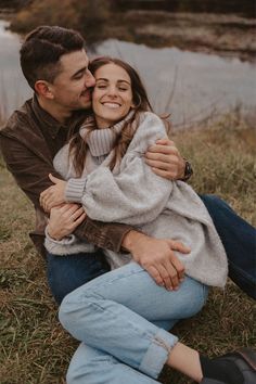 a man and woman sitting next to each other on the grass near water with trees in the background