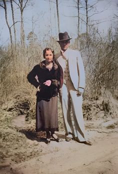 two people standing next to each other on a dirt road in front of some trees