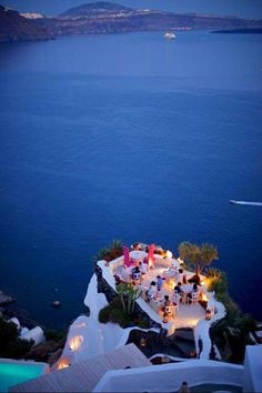 an outdoor dining area overlooking the ocean at night with lights on it and people sitting around