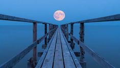 a full moon is seen over the water from a wooden pier at dusk with blue skies