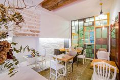 a dining room with white chairs and tables in front of a brick wall, surrounded by greenery