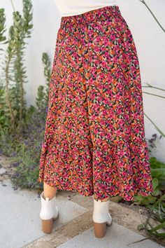 a woman in white shirt and colorful skirt standing on sidewalk next to planter with purple flowers