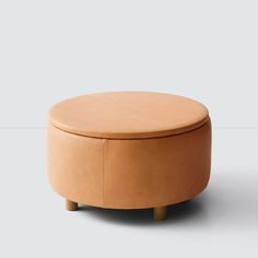 a small round ottoman with wooden legs and a tan leather cover on the top, sitting in front of a gray background