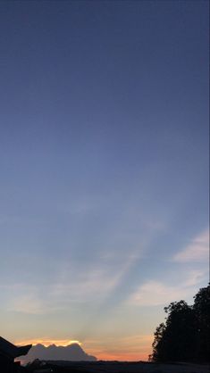 the sun is setting behind some trees and mountains in the distance, with an airplane flying low to the ground