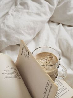 an open book sitting next to a cup of coffee on top of a bed with white sheets