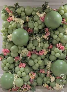 an arrangement of green balloons, flowers and greenery on a white wall with pink roses