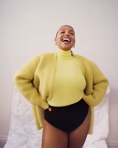 a woman in a yellow sweater and black shorts is laughing while standing on a bed