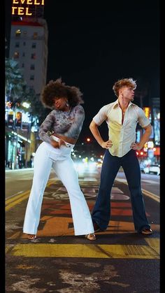 two people standing in the middle of a street at night with their hands on their hipss