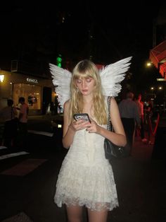 a woman dressed as an angel checks her cell phone in the street at night time