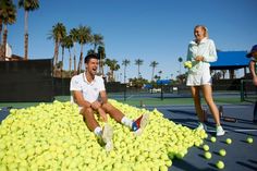 a man sitting on top of a pile of tennis balls next to a woman holding a racquet