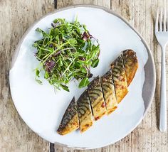 a white plate topped with fish next to a green leafy salad on top of a wooden table