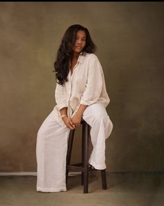 a woman sitting on top of a wooden chair next to a brown wall and wearing white pants