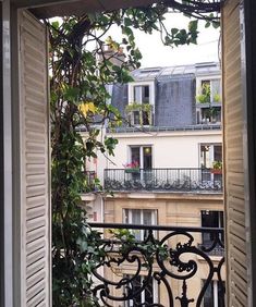 an open window looking out onto a balcony with plants growing on the railing and buildings in the background