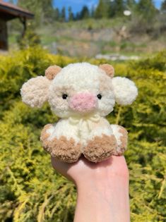 a hand holding a small stuffed animal in it's right hand with bushes and trees in the background