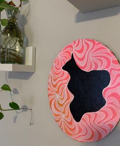 a pink and black circular painting on the wall next to a vase with green plants