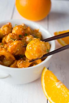 a white bowl filled with orange chicken next to an orange and two chopsticks