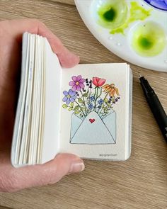 a person holding an open book with flowers in it and a pen on the table