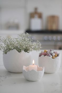 two white vases with flowers and rocks in them on a counter top next to a lit candle