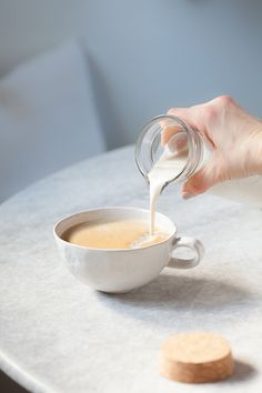 a person pouring milk into a cup on top of a white table next to a cookie