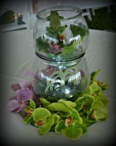 two glass vases filled with green and purple flowers on top of a white table