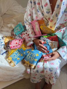 a woman sitting on a bed holding several bags of snacks and candy in her hands