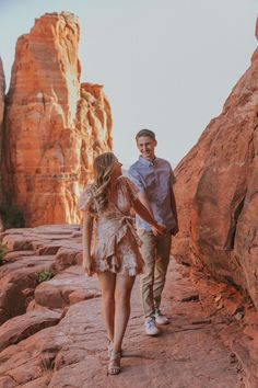 a man and woman holding hands while walking through the rocks in front of some mountains