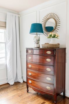 a wooden dresser with a blue lamp on top of it next to a white curtain