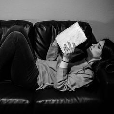 a woman laying on top of a black couch holding a book in her right hand