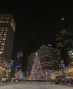 a christmas tree is lit up in the middle of an ice rink surrounded by tall buildings