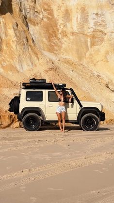 a woman standing next to a white jeep on top of a sandy beach in front of a cliff