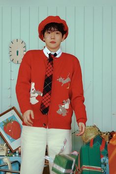 a young man in a red sweater and tie standing next to christmas presents with a clock on the wall behind him