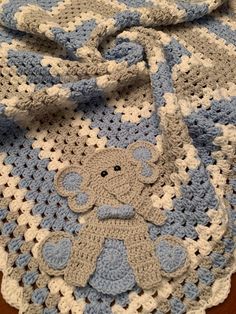 a blue and white crocheted blanket with a teddy bear on the bottom, sitting on a wooden floor