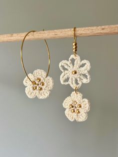 two crocheted flowers hang from gold hoop earrings on a wooden stick against a gray background