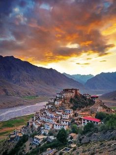 a village on top of a hill with mountains in the background and clouds above it