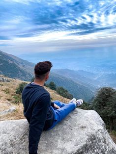 a man sitting on top of a large rock next to a valley and mountain range