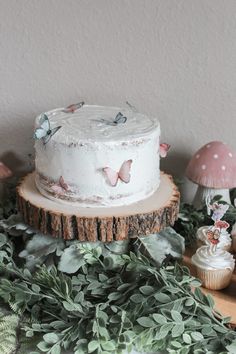 a white cake sitting on top of a wooden table next to cupcakes and plants