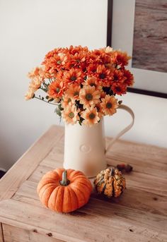 a white vase filled with orange flowers on top of a wooden table next to small pumpkins