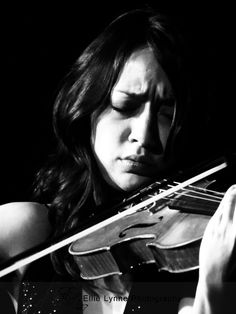 a woman holding a violin in her right hand and looking down at the camera with eyes closed