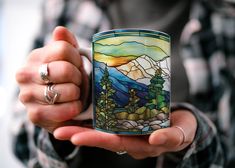 a person holding a stained glass mug with mountains in the background and trees on it