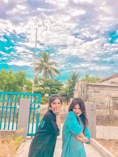 two women standing next to each other in front of a blue gate and sky with clouds